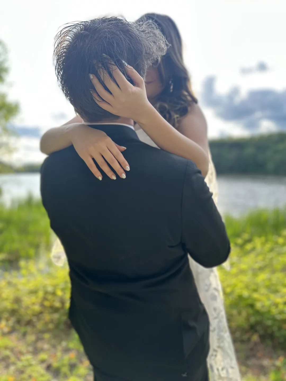 A couple holding each other close by a lake, her hands behind his head.