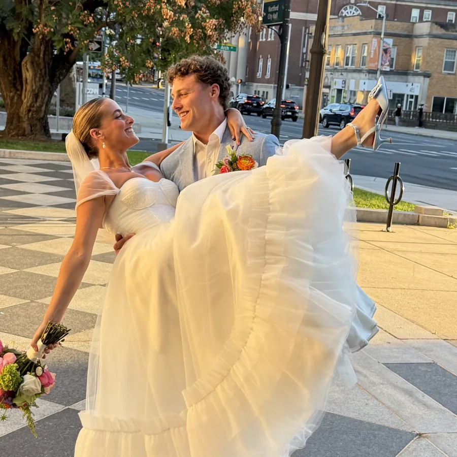 A groom carrying a bride in a tulle gown and veil on a city sidewalk, her bouquet in one hand