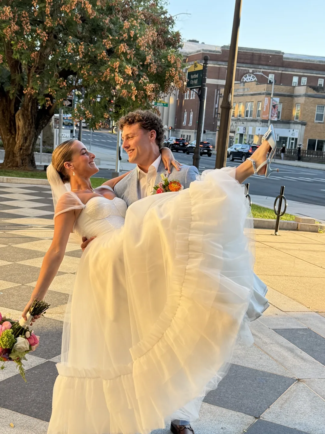 A groom carrying his bride along a city sidewalk, both of them laughing.
