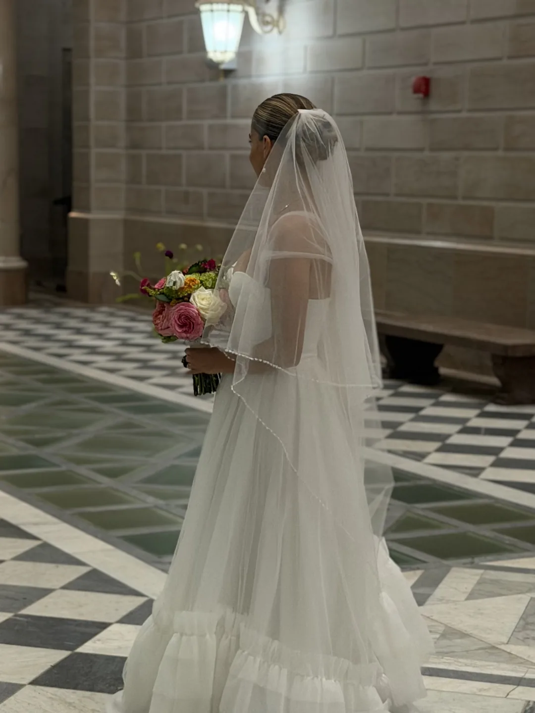 A bride from behind, holding her bouquet in a stone hallway with a checkered marble floor.